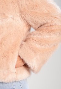 Ljust rosa fuskpälsjacka med ärmar, handen nedstoppad i sidofickan, visad tillsammans med ljusblå denimjeans på en neutral bakgrund.