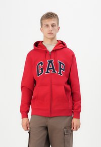 Rode rits-hoodie van katoenmix. Kenmerken een groot marineblauw "GAP" logo op de voorkant, een kangoeroezak en een capuchon met trekkoord.