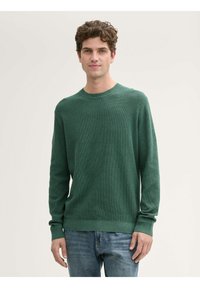 Maglione verde a maniche lunghe con un motivo di maglia testurizzato, scollatura rotonda e polsini a coste. Abbinato a jeans blu, mostrando un look casual.