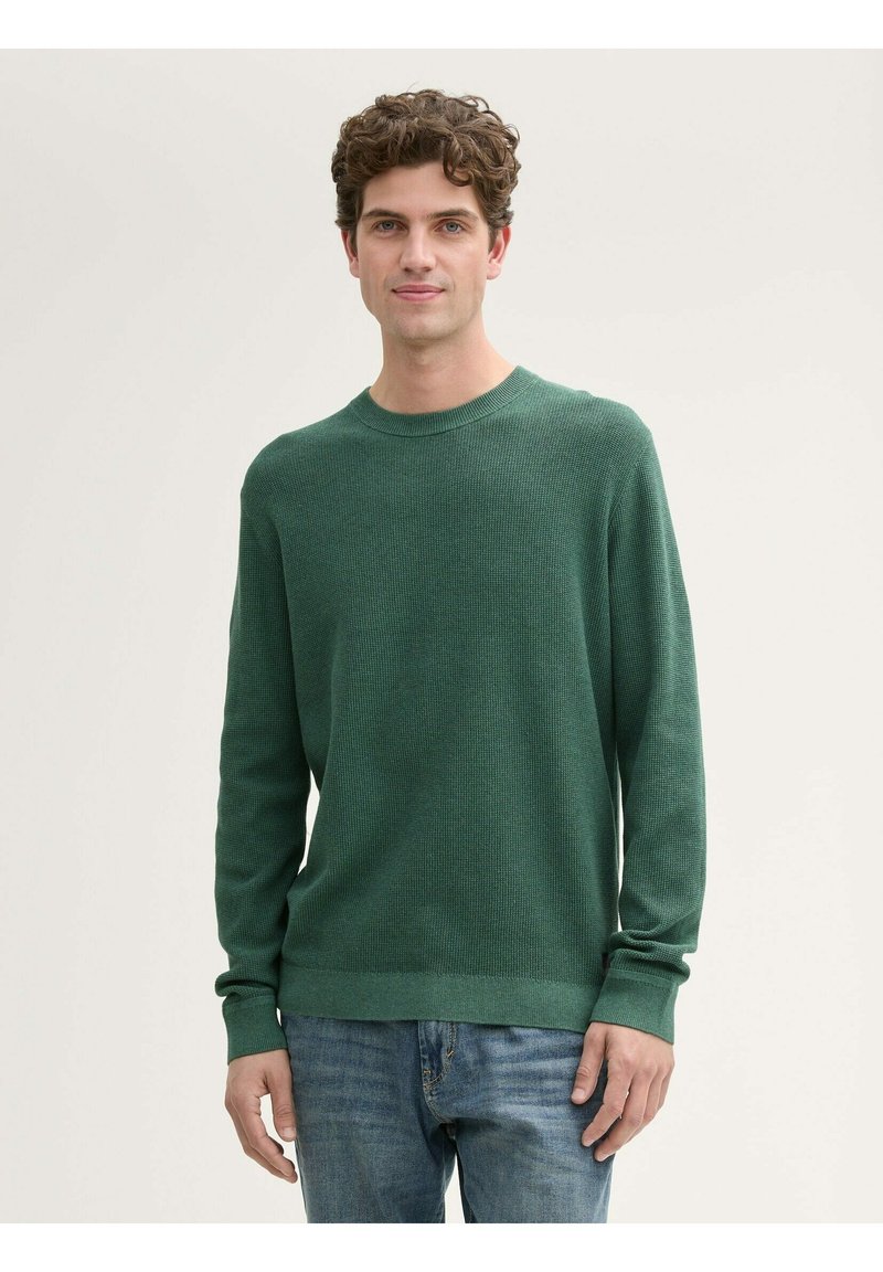 Maglione verde a maniche lunghe con un motivo di maglia testurizzato, scollatura rotonda e polsini a coste. Abbinato a jeans blu, mostrando un look casual.