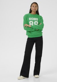 Zelená mikina s bílým potiskem "SOHO 88", kombinovaná s černými rozšířenými kalhotami a světlou obuví, na jednobarevném pozadí.
