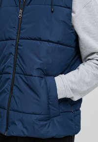 Tums zilas polsterēta veste ar priekša rāvējslēdzēju, sānu kabatām un pelēkajiem piedurkņu akcentiem. Gluda tekstūra ar quilted dizainu siltuma nodrošināšanai.