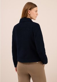 Kvinna i en marint blue texturerad jacka med krage och beige byxor, sedd bakifrån mot en enkel ljus bakgrund.