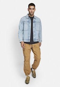 Light blue denim jacket with two front pockets, worn over a black shirt, paired with tan cargo pants and green sneakers with brown accents.