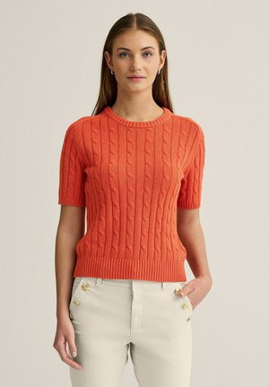 Ung kvinde med glat brunt hår iført kortærmet orange kabelstrikket sweater og off-white bukser med guldknapper, står mod en ensfarvet væg.