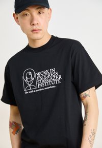 T-shirt de algodão preta com uma graphic frontal com o texto "INÍCIO DE TRABALHO INSTITUTO STARGAZER" em branco. Corte relaxado e mangas curtas.