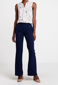 Kvinna iklädd ärmlös vit blommig blus, marinblå vida byxor med knapprader, och beige klackskor med blockklack, stående mot vit bakgrund.