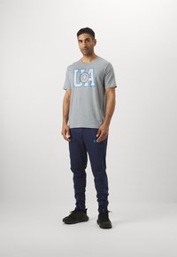 Grå bomulds t-shirt med blå "UA" grafik, parret med marineblå sweatpants med blå sidestriber og sorte træningssko.
