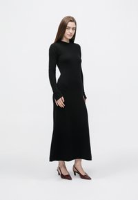 Długa czarna sukienka dziana o dopasowanym kroju, z długimi rękawami i zaokrąglonym dołem. Noszona z bordowymi butami na obcasie. Gładka faktura.