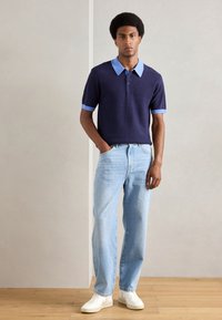 Marinblå pikétröja med ljusblå krage och ärmslut, i kombination med ljusblå jeans. Modellen står med händerna i fickorna och har vita sneakers.