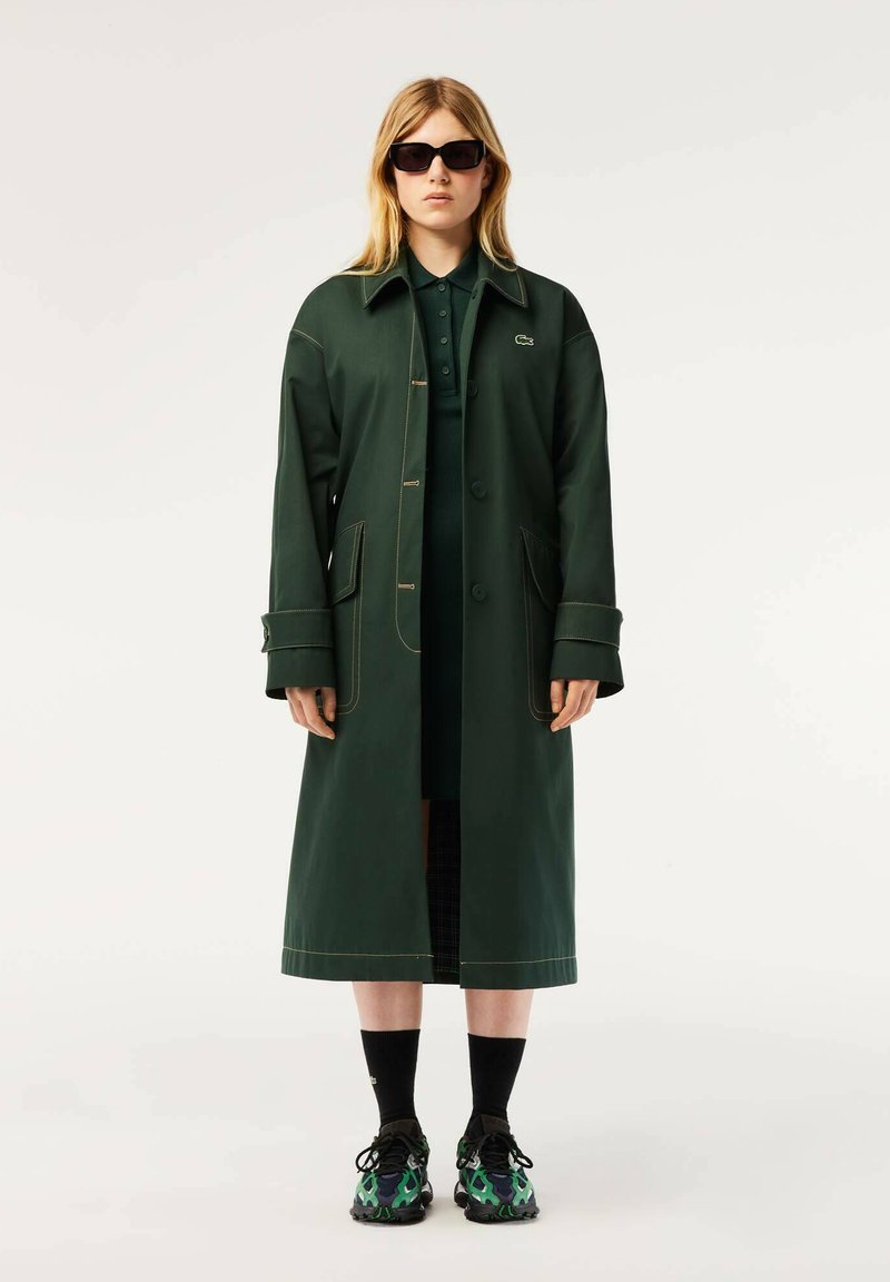 Manteau vert, longueur genoux, avec une fermeture à boutons, de grandes poches et des coutures jaunes contrastantes, porté sur un polo vert foncé et une jupe à motifs.