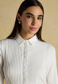 White blouse featuring a pleated texture on the front, pointed collar, and button closure. Model wears gold drop earrings.