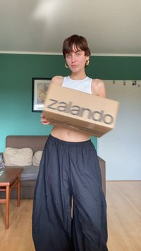 Hnědá kartonová krabice s nápisem "Zalando" vytištěným tučným písmem, držena před osobou v bílém tílku a černých širokých kalhotách.