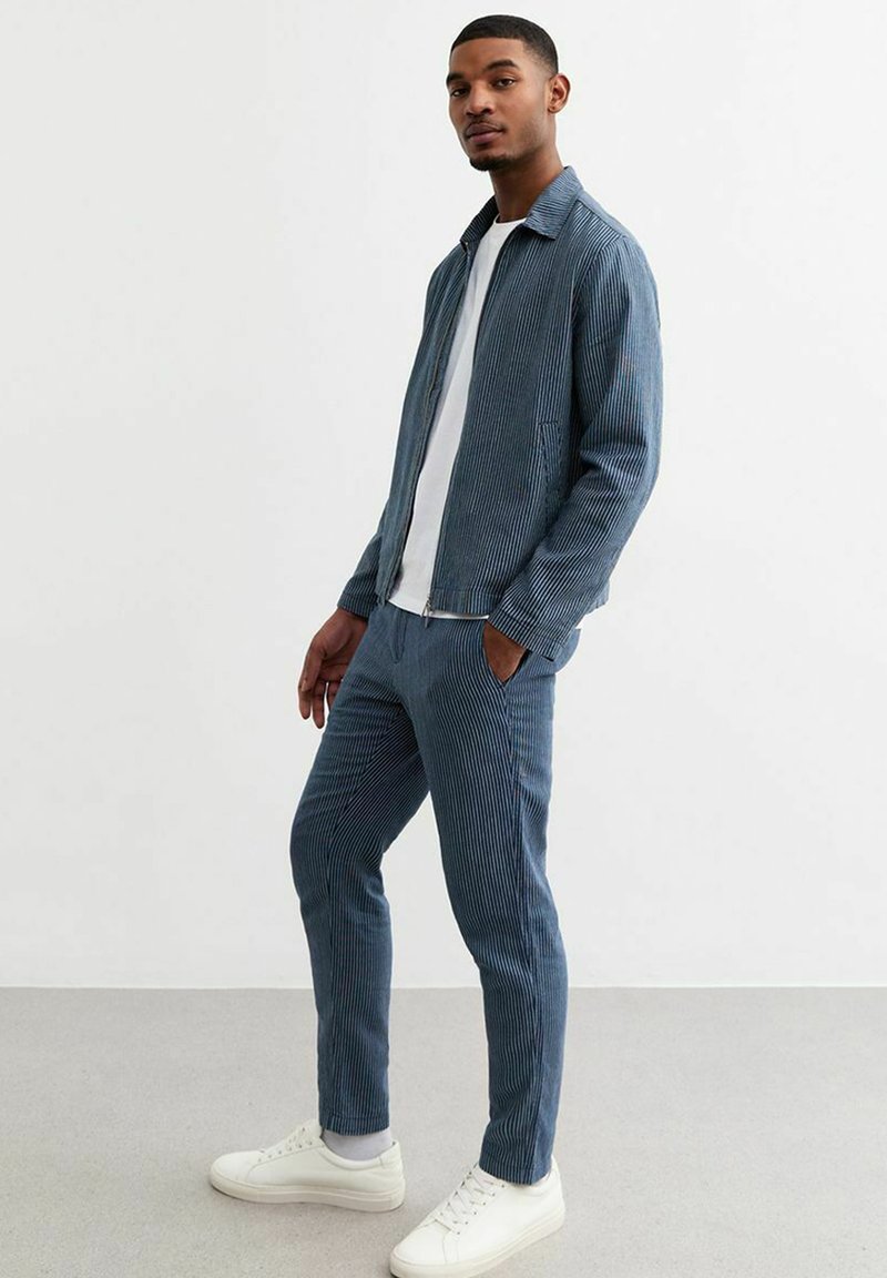 Blauwe corduroy-outfit bestaande uit een jas met rits en slim-fit broeken, gecombineerd met witte sneakers, met verticale strepen.