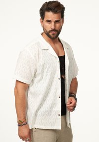 Kurzärmliges weißes Hemd mit einem strukturierten Blumenmuster, kombiniert mit einem schwarzen Tanktop und beigen Hosen, accessorisiert mit Armbändern.