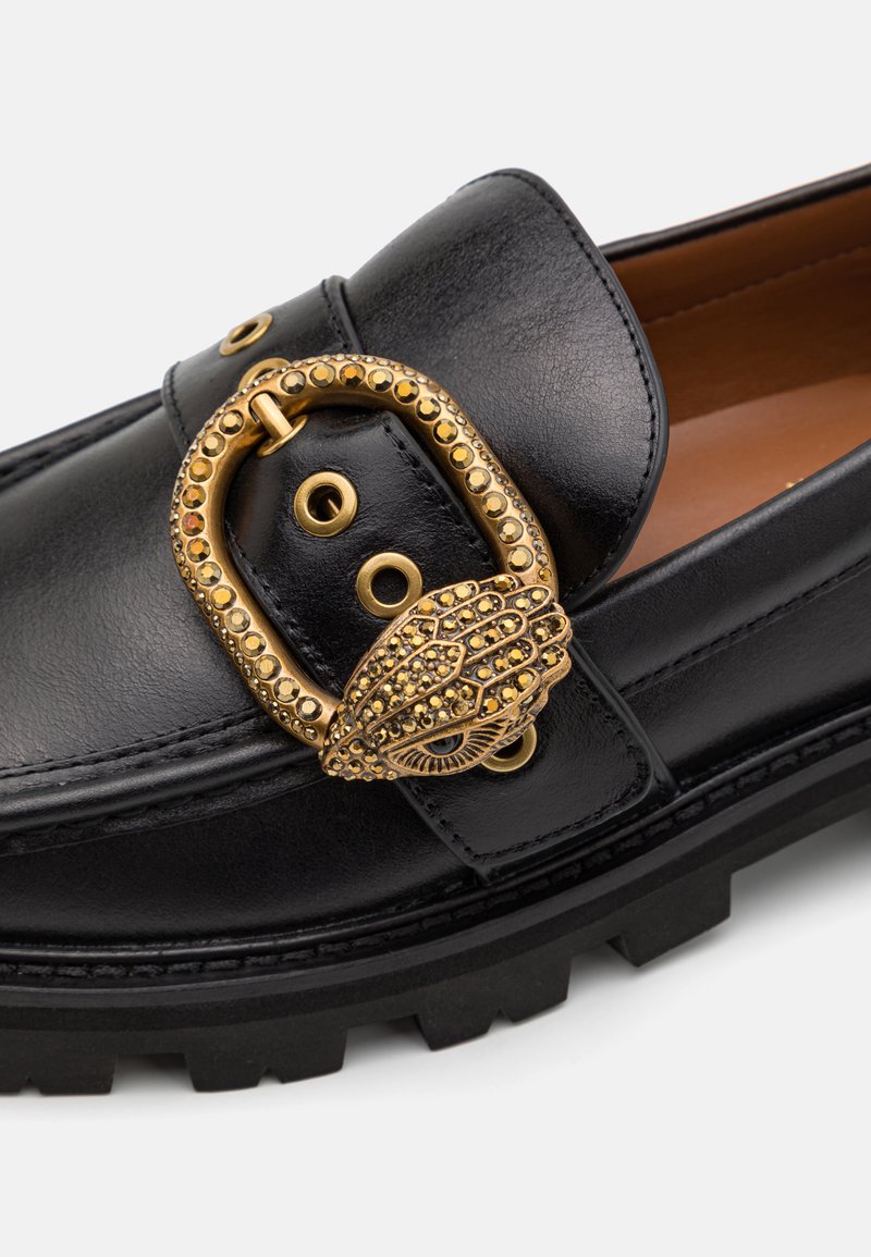 Mocassins en cuir noir avec une boucle décorative en or ornée de strass et de découpes ; surface texturée, bout arrondi et semelle épaisse.