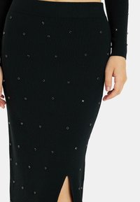 Falda de punto negra con canalé y abertura lateral, adornada con acentos metálicos dispersos, que exhibe un diseño ceñido y una textura suave.