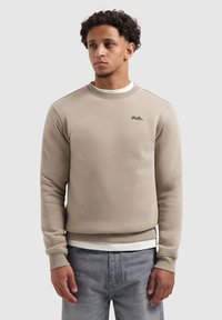 Man met krullend haar in een beige sweatshirt met het label "Ballin" over een wit shirt en grijze jeans, staande tegen een effen lichte achtergrond.