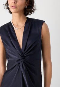 Meripäivän satiinimekko, jossa on syvä V-pääntie ja kierrerintamuotoilu. Omaavat hihattoman muotoilun ja sileän tekstuurin, yhdistettynä helmikaulaketjuun.