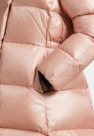 Mão parcialmente dentro do bolso de uma jaqueta de inverno acolchoada, brilhante e de cor rosa claro, com costura e enchimento visíveis.