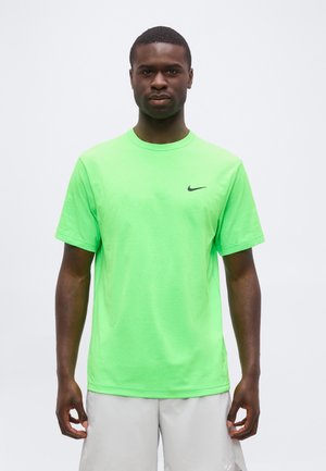 Mężczyzna stojący przodem, ubrany w jasnozielony t-shirt Nike i jasnoszorty na jednolitym tle.