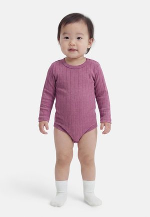 Tout-petit debout, de face, portant un body côtelé à manches longues rose et des chaussettes blanches sur un fond blanc uni.