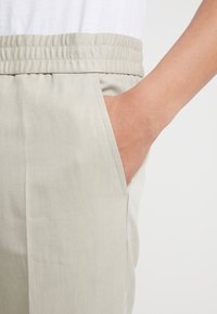 Light beige pants featuring an elastic waistband and side pocket, made of a smooth, textured fabric with a subtle diagonal pattern.