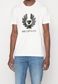 Camiseta de algodón blanca con un gran logo de águila negra con un emblema central, y el texto "BELSTAFF" debajo en una fuente negra en negrita. Corte casual.