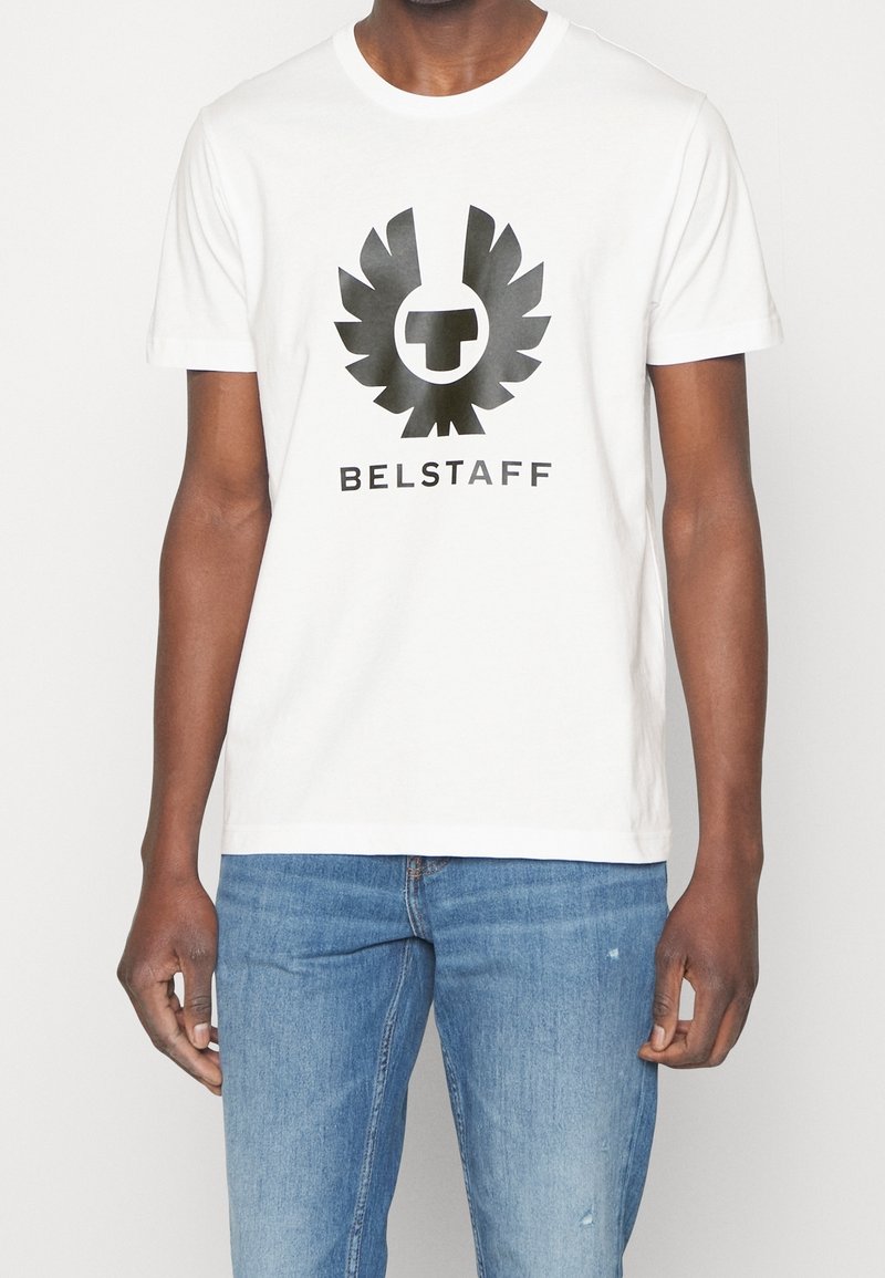 Camiseta de algodón blanca con un gran logo de águila negra con un emblema central, y el texto "BELSTAFF" debajo en una fuente negra en negrita. Corte casual.