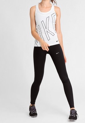 Kobieta w białym topie Nike i czarnych legginsach Nike stoi z rozstawionymi nogami na jednolitym szarym tle.