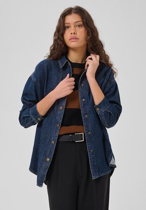 Femme aux longs cheveux bouclés portant une veste en jean déboutonnée sur un pull rayé marron et noir et un pantalon noir avec une ceinture.