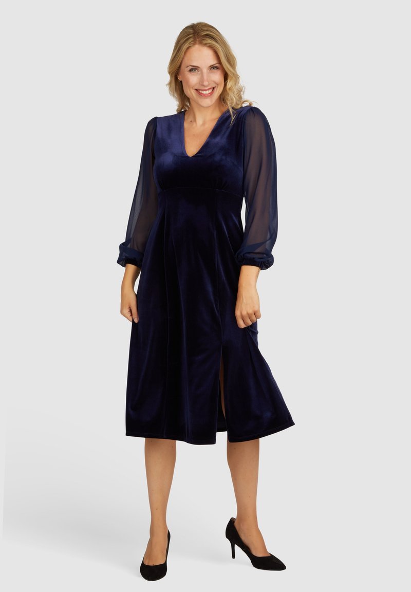 Navy-blaues Samtkleid mit transparenten Puffärmeln, V-Ausschnitt und Vorder-Schlitzen; der taillierte Oberteil geht in einen ausgestellten Rock über. Schwarze High Heels.