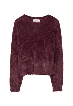 Person wearing dark flared jeans, a fluffy dark purple sweater over a white shirt, and black shoes standing against a pink background.