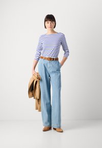 Striped langermet skjorte i hvit og blå, sammen med lyseblå vidde jeans, brun belte, og beige jakke som holdes i hånden. Brune sko.