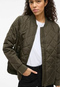 Oljčno zelena prešita bomber jakna z zadrgo spredaj, rebrastimi manšetami in belo majico pod njo, kombinirana s črnimi hlačami.