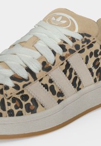 Μπεζ αθλητικό παπούτσι Adidas με λευκά κορδόνια και τρεις ανοιχτό μπεζ ρίγες πάνω σε μαύρο και καφέ λεοπάρ μοτίβο.