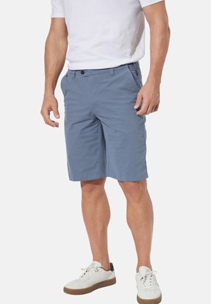 Mann trägt blaue knielange Shorts, weißes T-Shirt und weiße Turnschuhe und steht mit locker an den Seiten hängenden Händen.