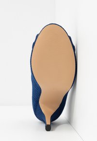 Chaussure à talon haut bleue texturée vue par la semelle, avec une semelle beige lisse et une petite pointe de talon noire.