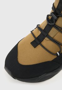 Καφέ και μαύρο αθλητικό παπούτσι με υφή στην επάνω πλευρά, ελαστικά κορδόνια και ενισχυμένη μύτη. Η σόλα έχει μιαdistinct καμπύλη μορφή.