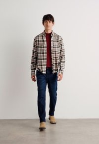 Checkered shirt in beige, green, and red tones, paired with a maroon sweater and dark blue jeans. Brown ankle boots complete the outfit.
