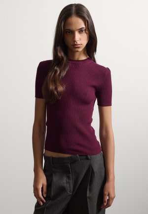Joven mujer con cabello largo y oscuro que lleva una camiseta burdeos de manga corta con textura acanalada y pantalones gris oscuro de talle alto sobre un fondo liso.