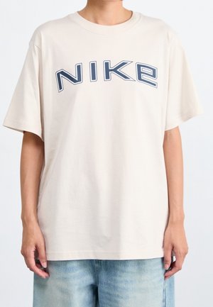 Osoba nosząca luźną jasnobeżową koszulkę z "NIKE" dużymi granatowymi literami oraz jasnoniebieskie szorty jeansowe.