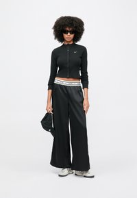 Μαύρο ριμπ μακρυμάνικο τοπ με κουμπιά, μαύρο παντελόνι φαρδιάς γραμμής με λευκή ταινία, μαύρο καπέλο και γκρι παπούτσια με μαύρες λεπτομέρειες.