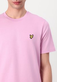 Rosa kortärmad t-shirt i bomull, med en liten broderad fågel-logotyp i svart och gult på vänster bröst.