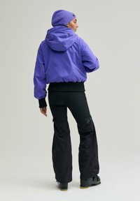 Person steht, trägt eine lilafarbene Kapuzenjacke, eine passende lilafarbene Mütze, schwarze Cargo-Hosen und schwarze Winterstiefel vor einem einfarbigen Hintergrund.