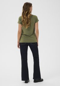 T-shirt verde oliva a maniche corte con una vestibilità comoda, abbinato a jeans svasati blu scuro, su uno sfondo bianco semplice.