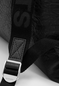 Detalle de un bolso de cuero negro que presenta una superficie texturizada, una correa plana con un logo y una hebilla de metal plateado con marca en relieve.