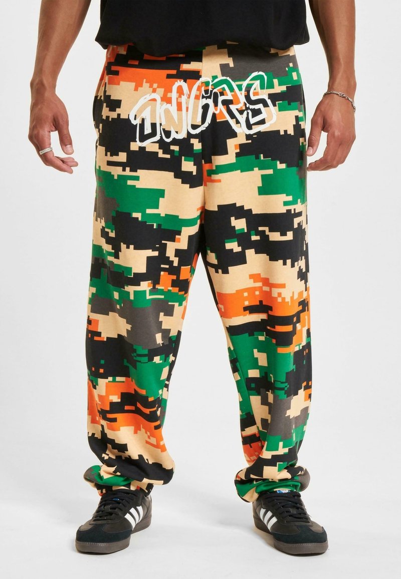 Mand iført løse bukser med digitalt camouflage-mønster i grønt, orange, sort og beige med teksten "SNIP" nær taljen og sorte sneakers med hvide striber.