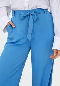 Pantalon large bleu en tissu texturé, avec une ceinture à nouer assortie à la taille et des poches latérales. Porté avec une chemise blanche à col.