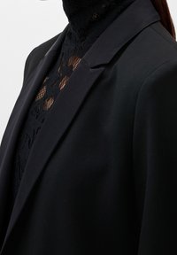 Blazer nero con una texture liscia, dotato di risvolto a scialle e spalle strutturate, indossato sopra un dolcevita in pizzo nero.